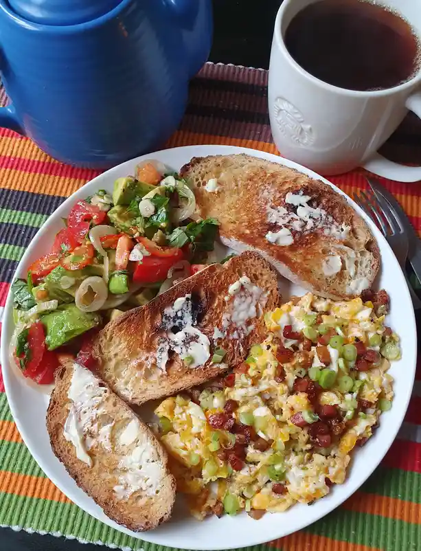 A plate of breakfast food including scrambled eggs with bacon and green onions, avocado tomato salad, and three slices of…