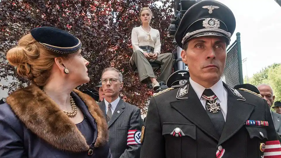 A woman in a fur-collared coat and hat speaks to a stern-looking man in a Nazi uniform adorned with insignia. Behind them, a…