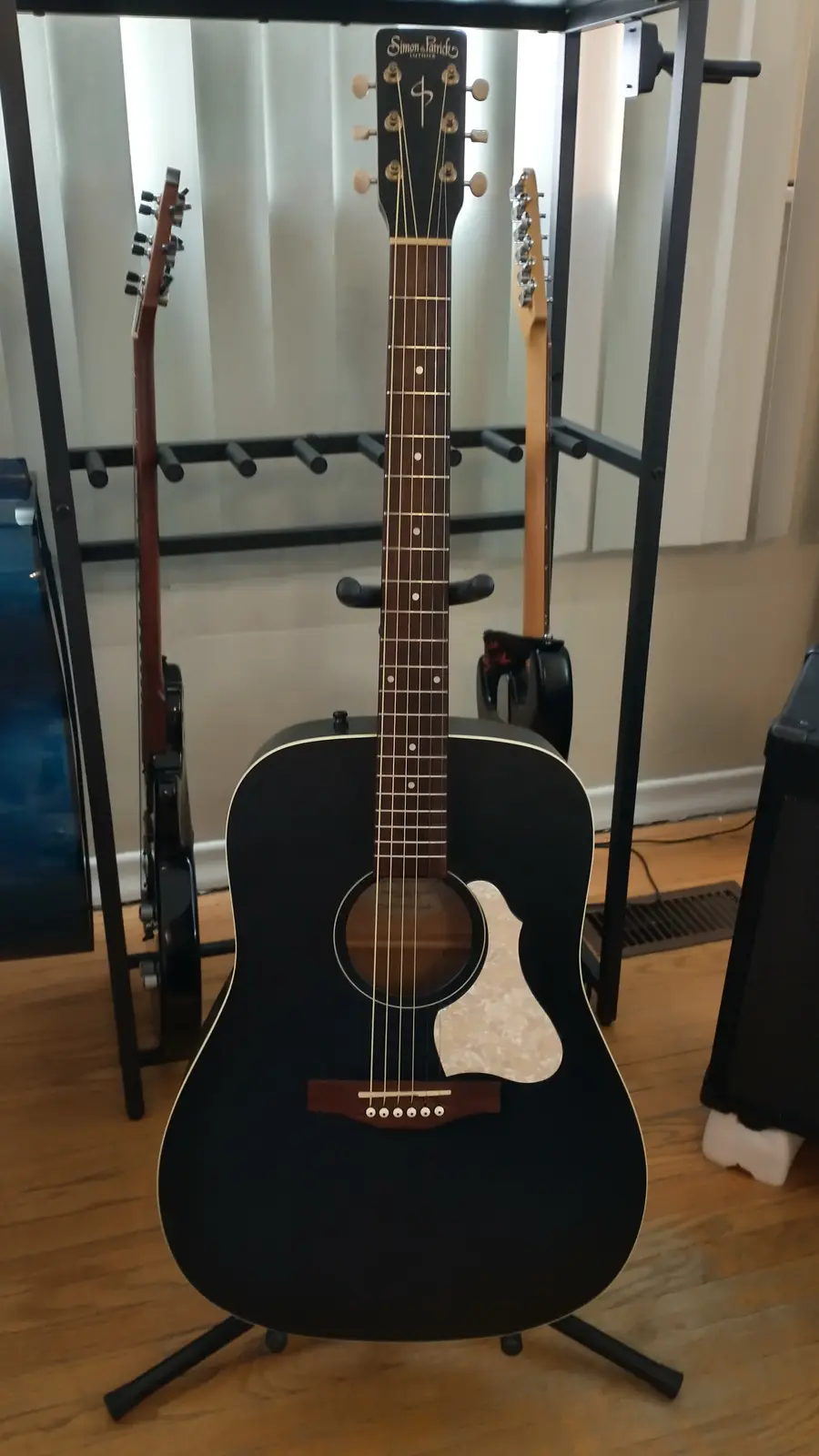 A black acoustic guitar with a white pickguard and 'Simon & Patrick Luthier' branding on the headstock stands upright on a…