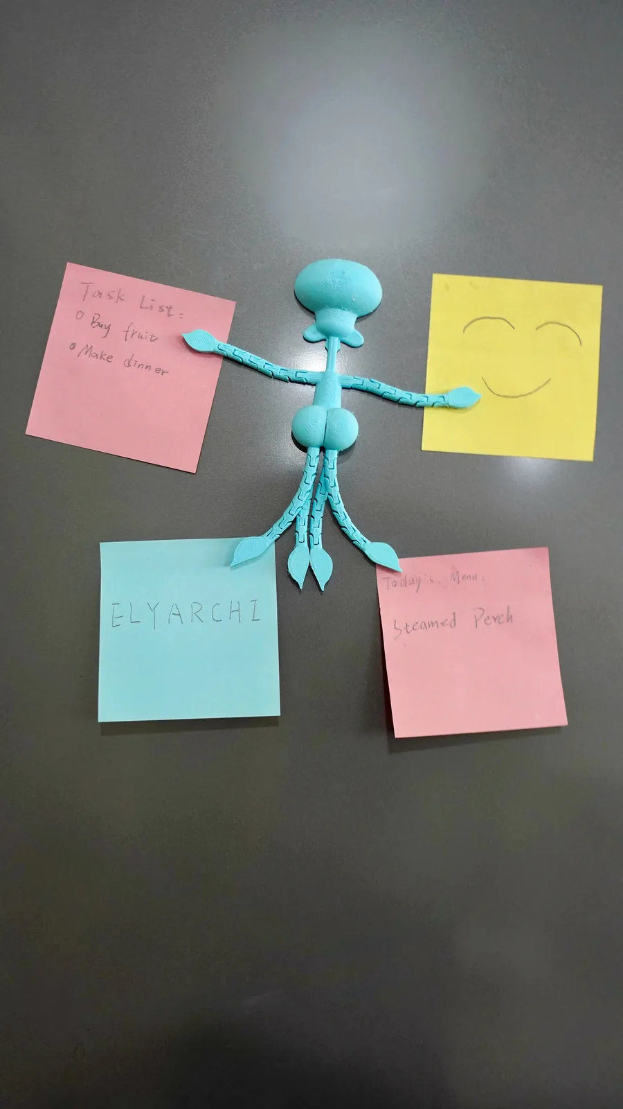 A light blue 3D-printed Squidward-like figurine is pinned to a dark surface with four sticky notes. The pink note on the…
