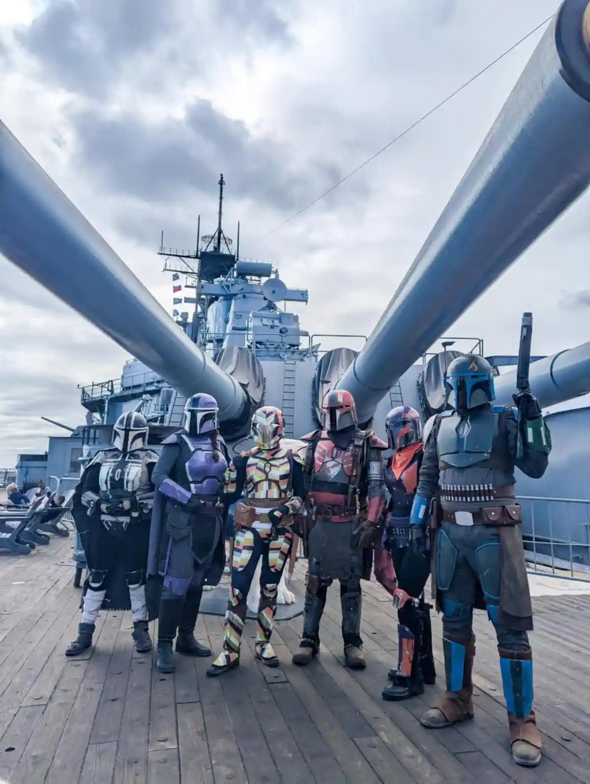 Six people dressed as Mandalorians from Star Wars stand on the deck of a large naval warship, framed by two massive gun…