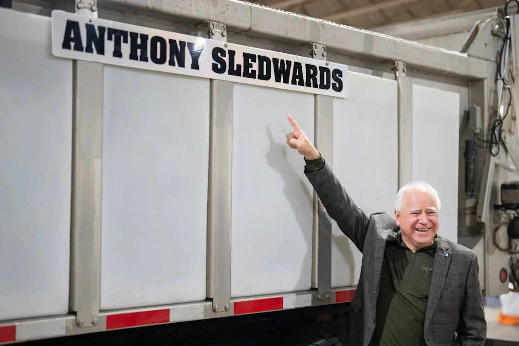 A smiling older man in a suit jacket points upward with his right index finger toward a white sign on a large truck that…