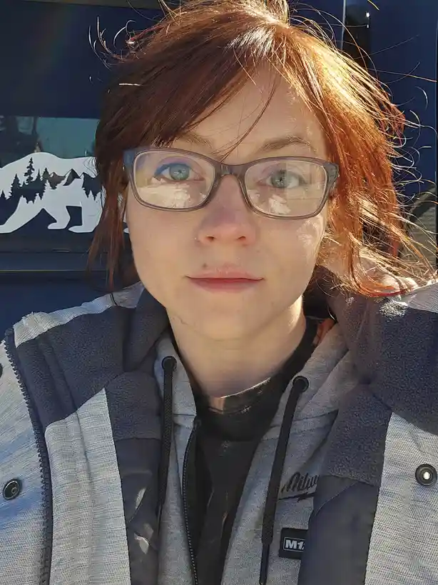 A person with red hair and glasses stares directly at the camera with a neutral expression, standing in front of a blue…