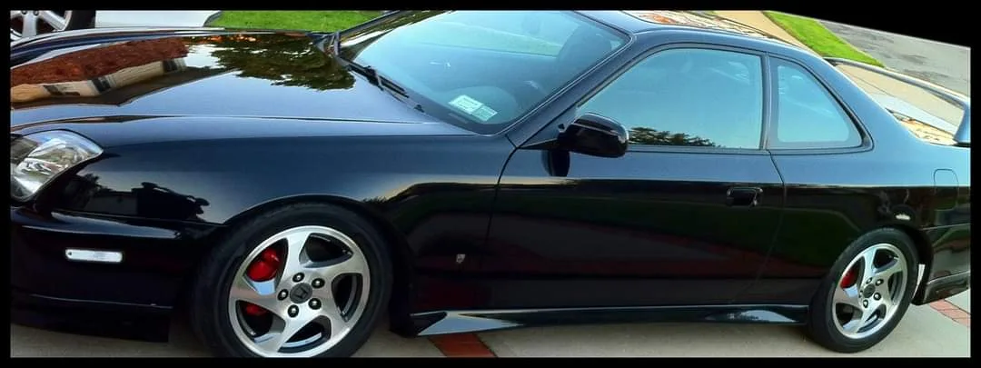 a glossy black nissan skyline r34 coupe is parked on a driveway, showing its side profile with silver multi-spoke wheels…