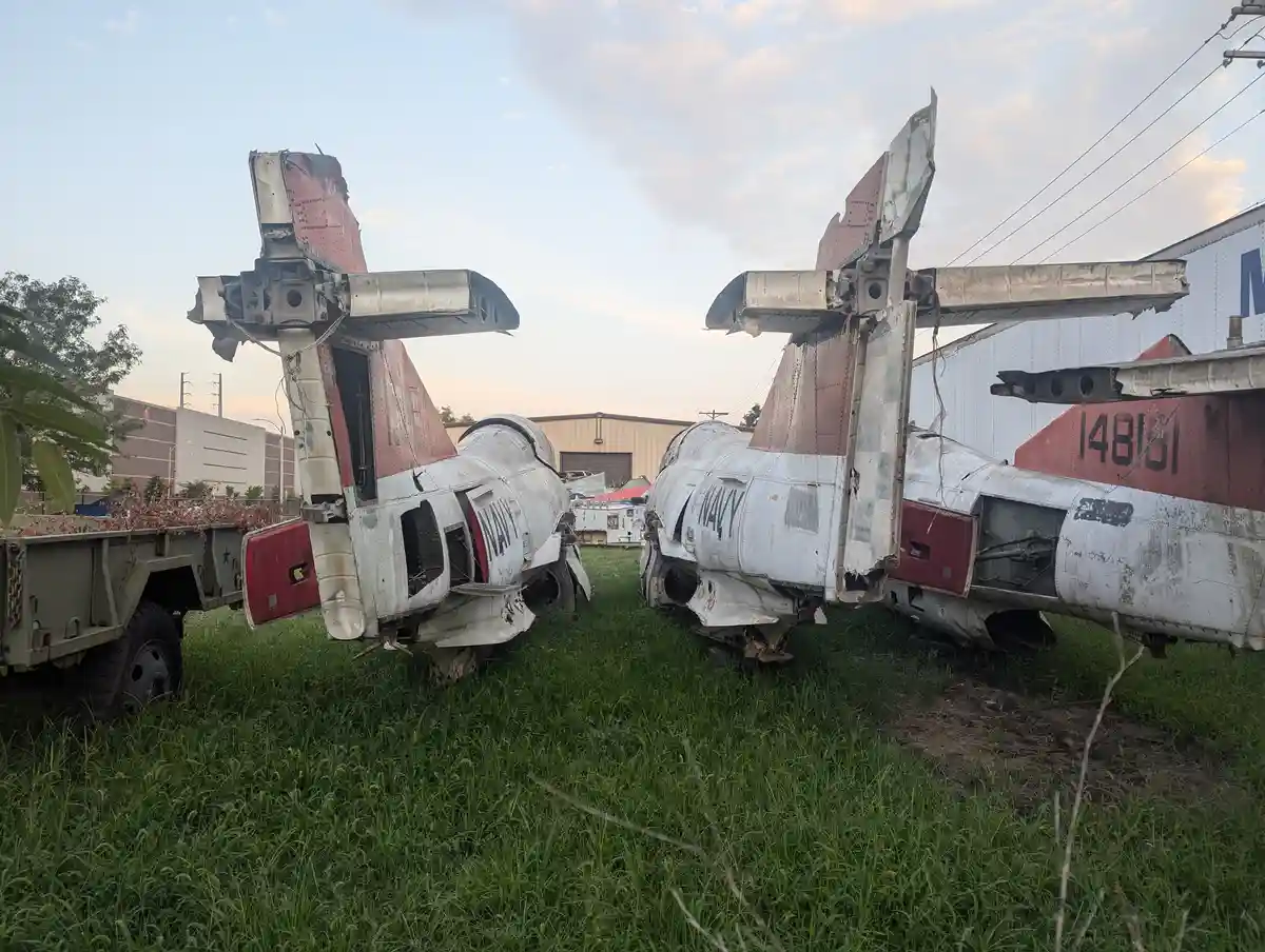Two heavily damaged, rusted, and stripped-down U.S. Navy aircraft fuselages lie abandoned in tall grass, their tails and…