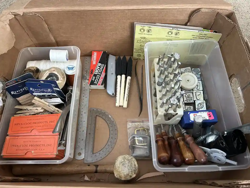 An overhead shot of a cardboard box filled with various crafting and leatherworking tools, including rulers, punches…