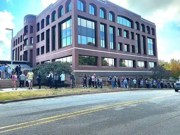 A long line of people stretches along the sidewalk in front of a large brick courthouse under a partly cloudy sky. The…
