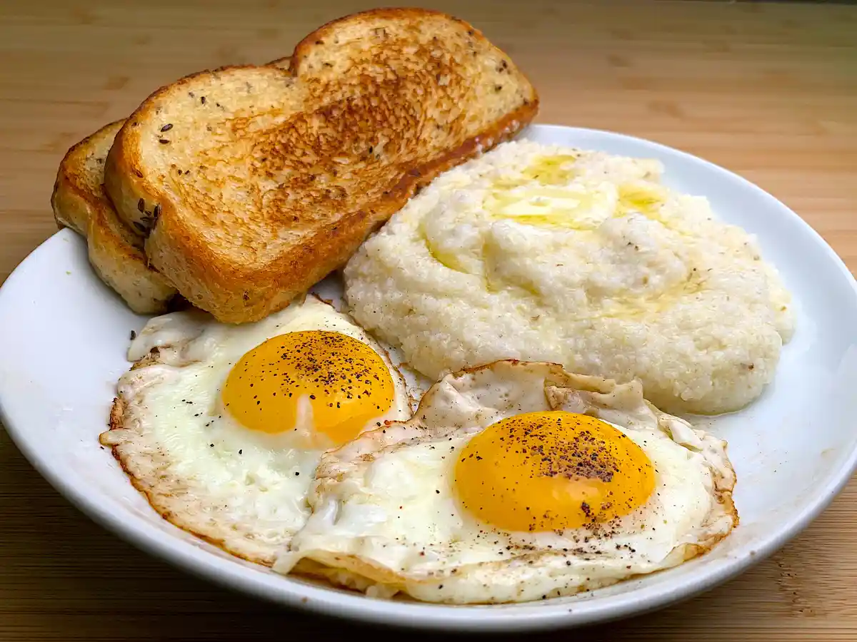 A white plate holds two sunny-side-up eggs seasoned with black pepper, a serving of creamy grits with melted butter, and…