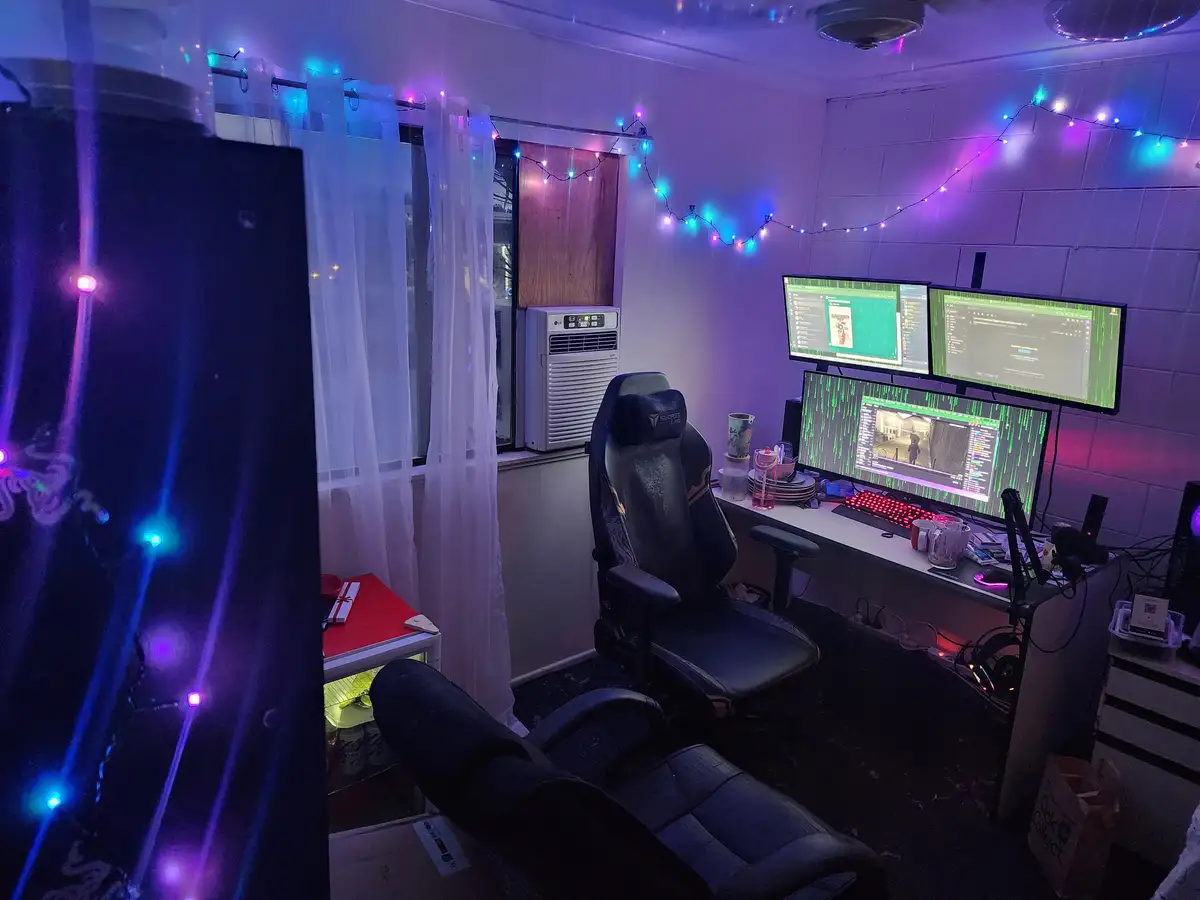 A dimly lit room with purple and blue string lights draped across the walls and curtains, featuring a gaming setup with…