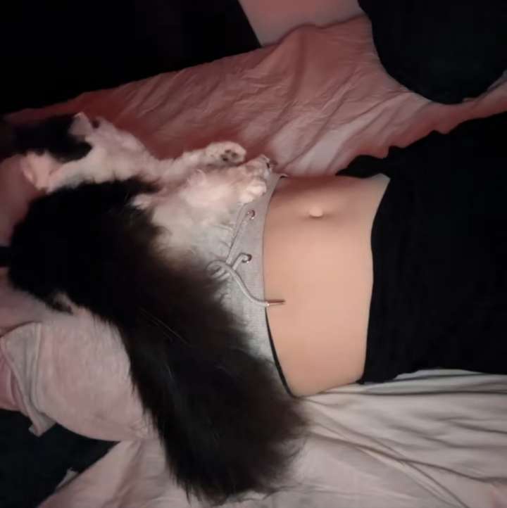 A person lies on their back in bed, wearing a grey top with a visible belly button and black bottoms, with a black-and-white…