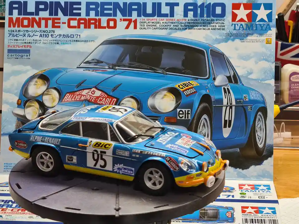 A blue 1:24 scale Alpine Renault A110 Monte Carlo '71 model car, number 95, sits on a display stand in front of its Tamiya…