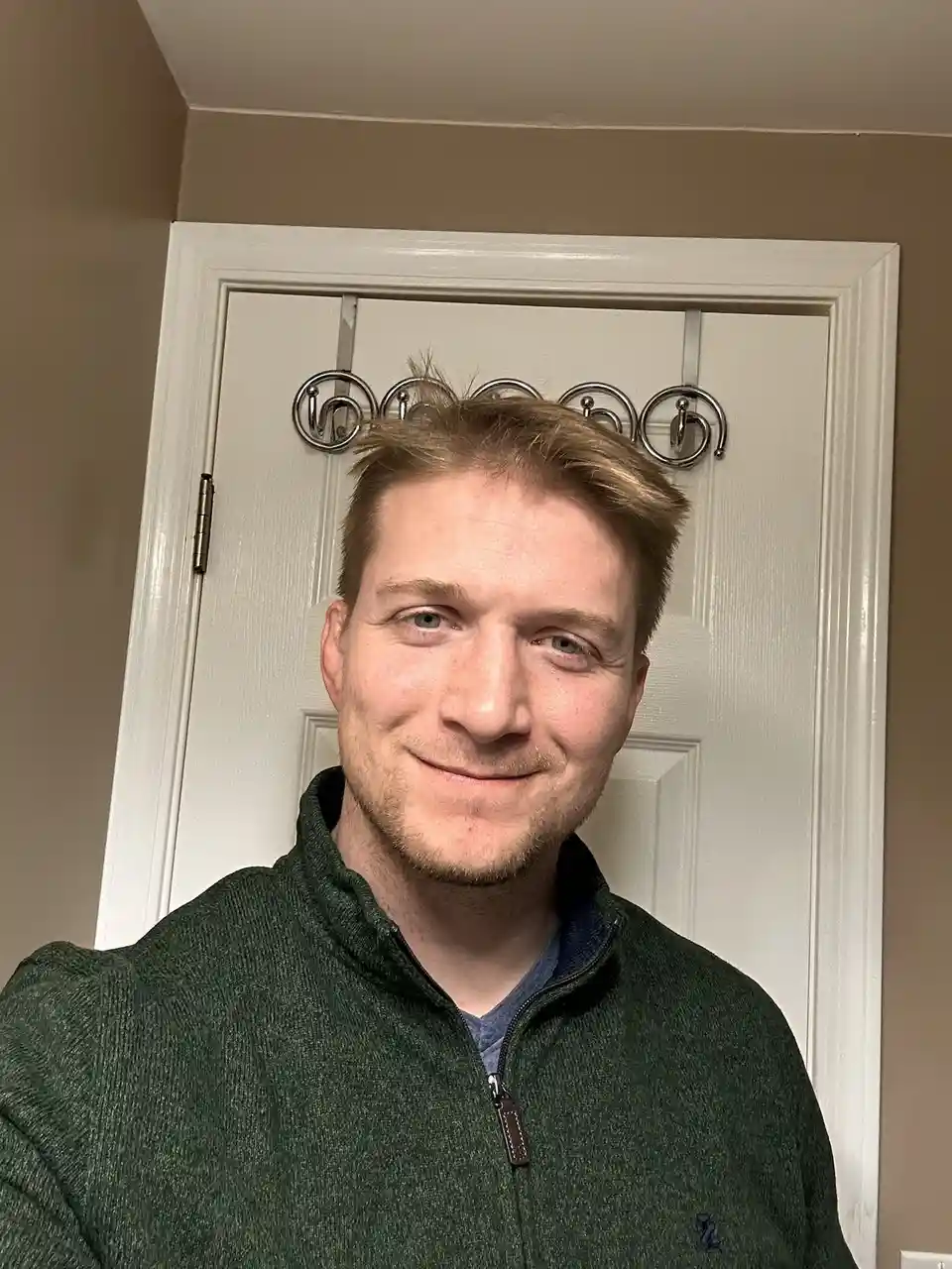 A man with light brown hair and a slight smile stands in front of a white door with a silver hook rack above his head. His…