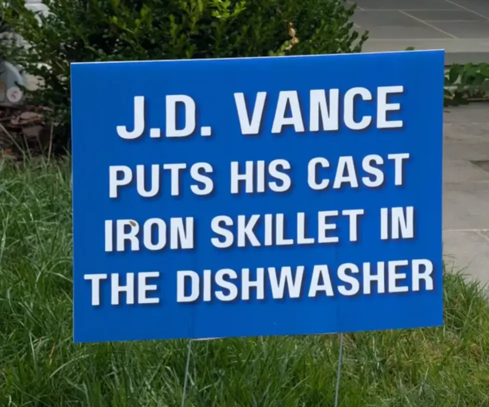 A blue political yard sign with white text stands in grass, reading 'J.D. VANCE PUTS HIS CAST IRON SKILLET IN THE…