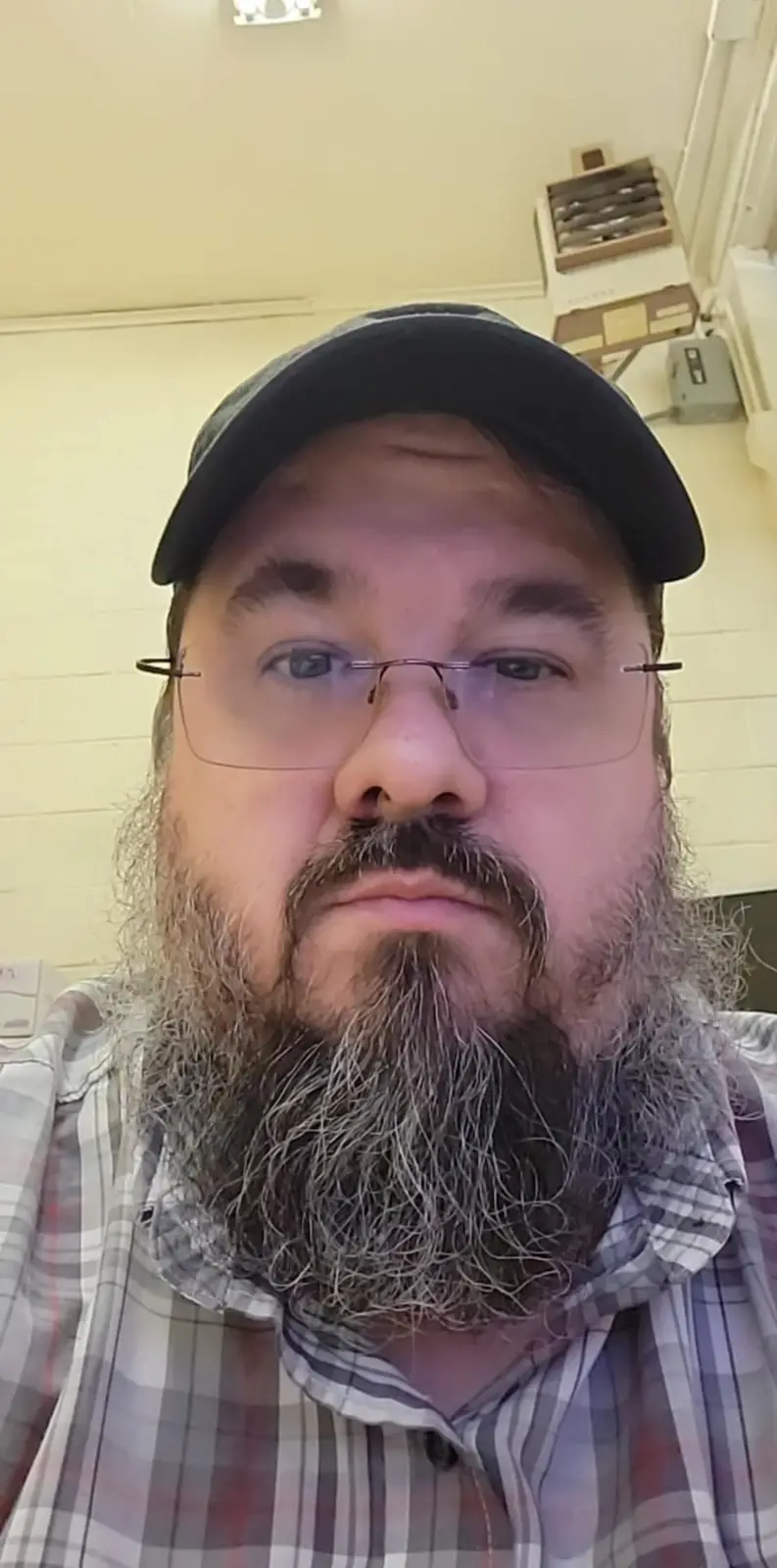 A man with a long, graying beard, wearing glasses and a black baseball cap, stares directly at the camera with a stern…