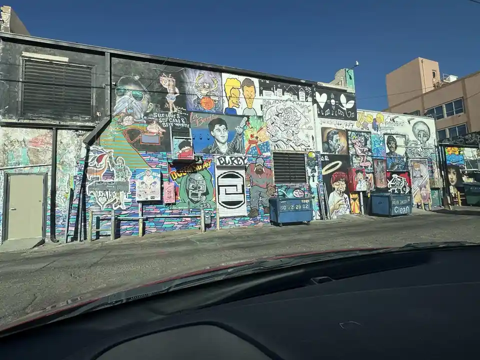 A photo taken from inside a car shows a long wall covered in colorful, eclectic street art and murals featuring cartoon…