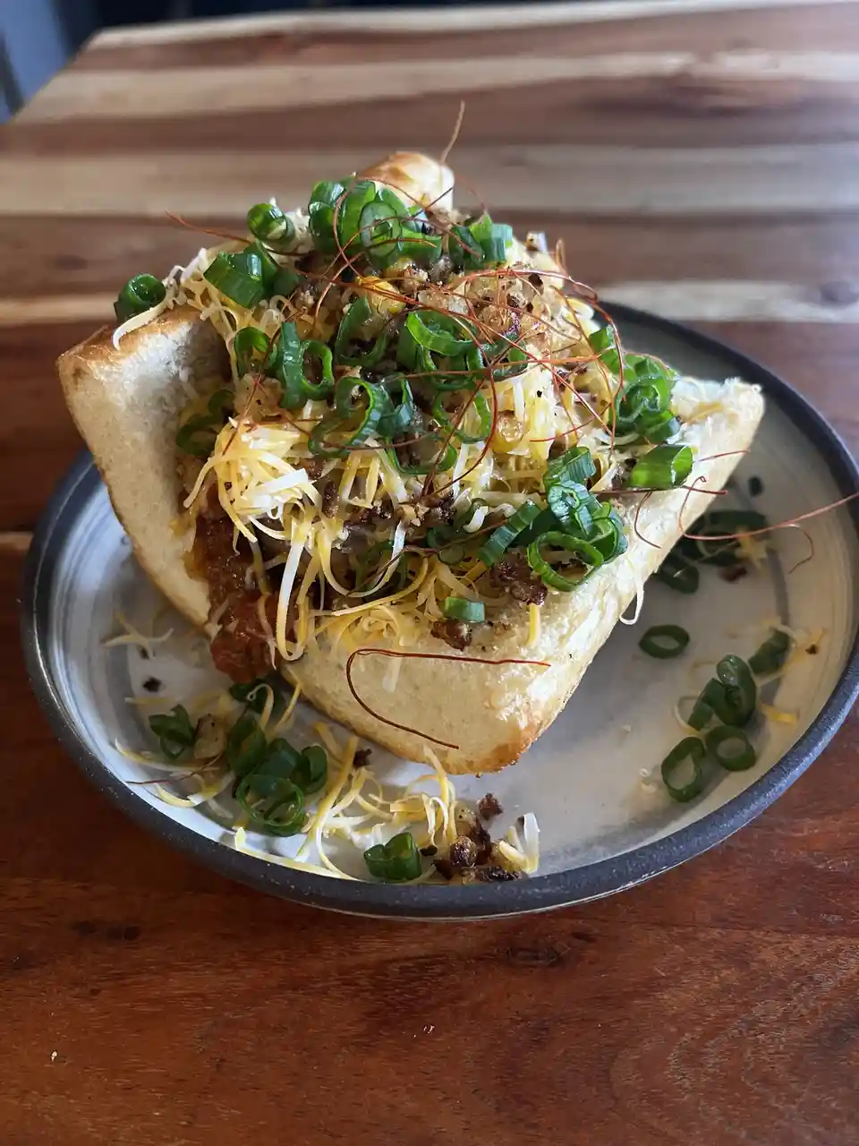 A close-up photo shows a chili dog served in a toasted bun on a ceramic plate, topped with shredded cheese, chopped green…
