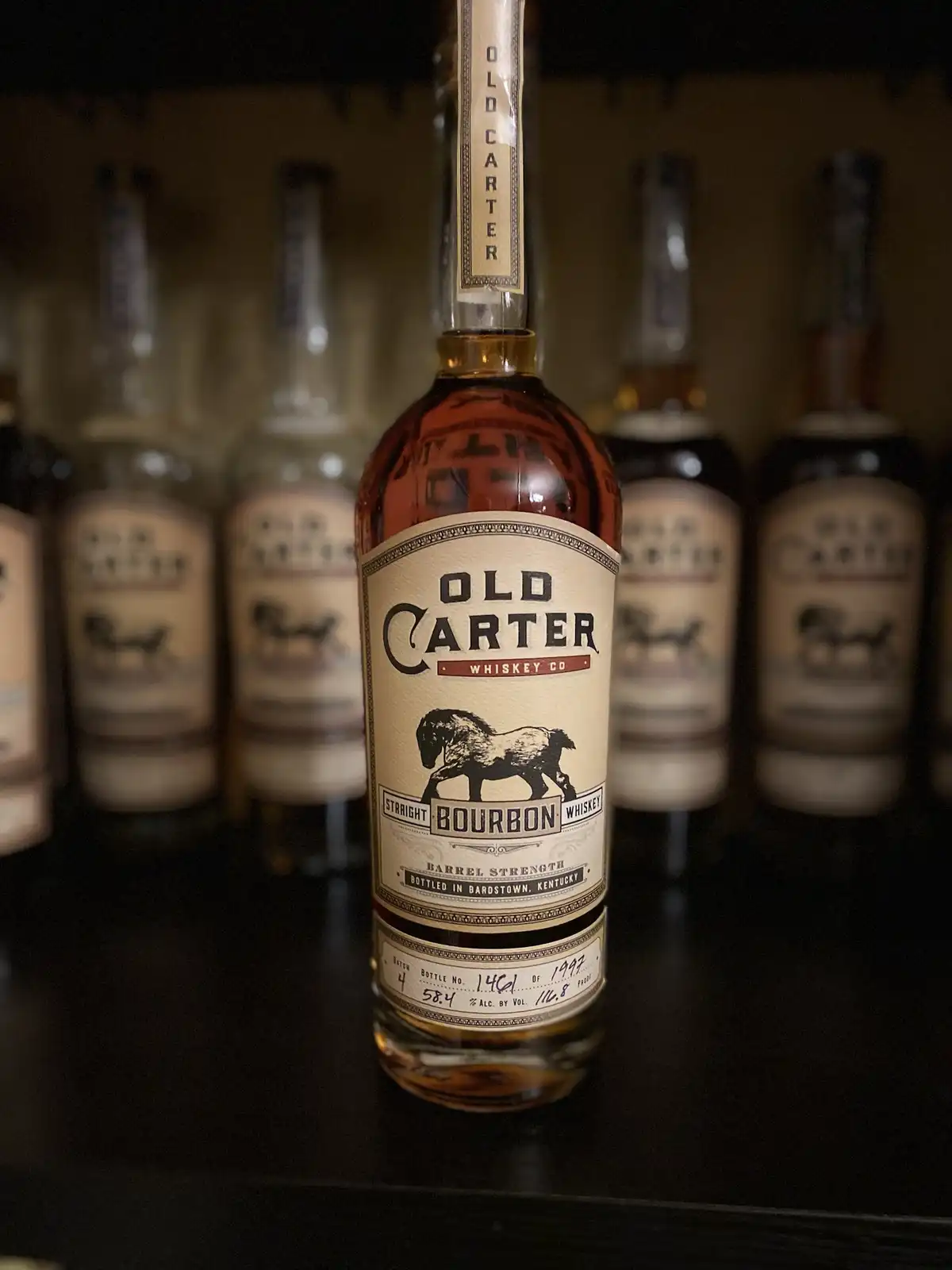 A close-up photograph of a bottle of Old Carter Straight Bourbon Whiskey, prominently displayed on a shelf with several…