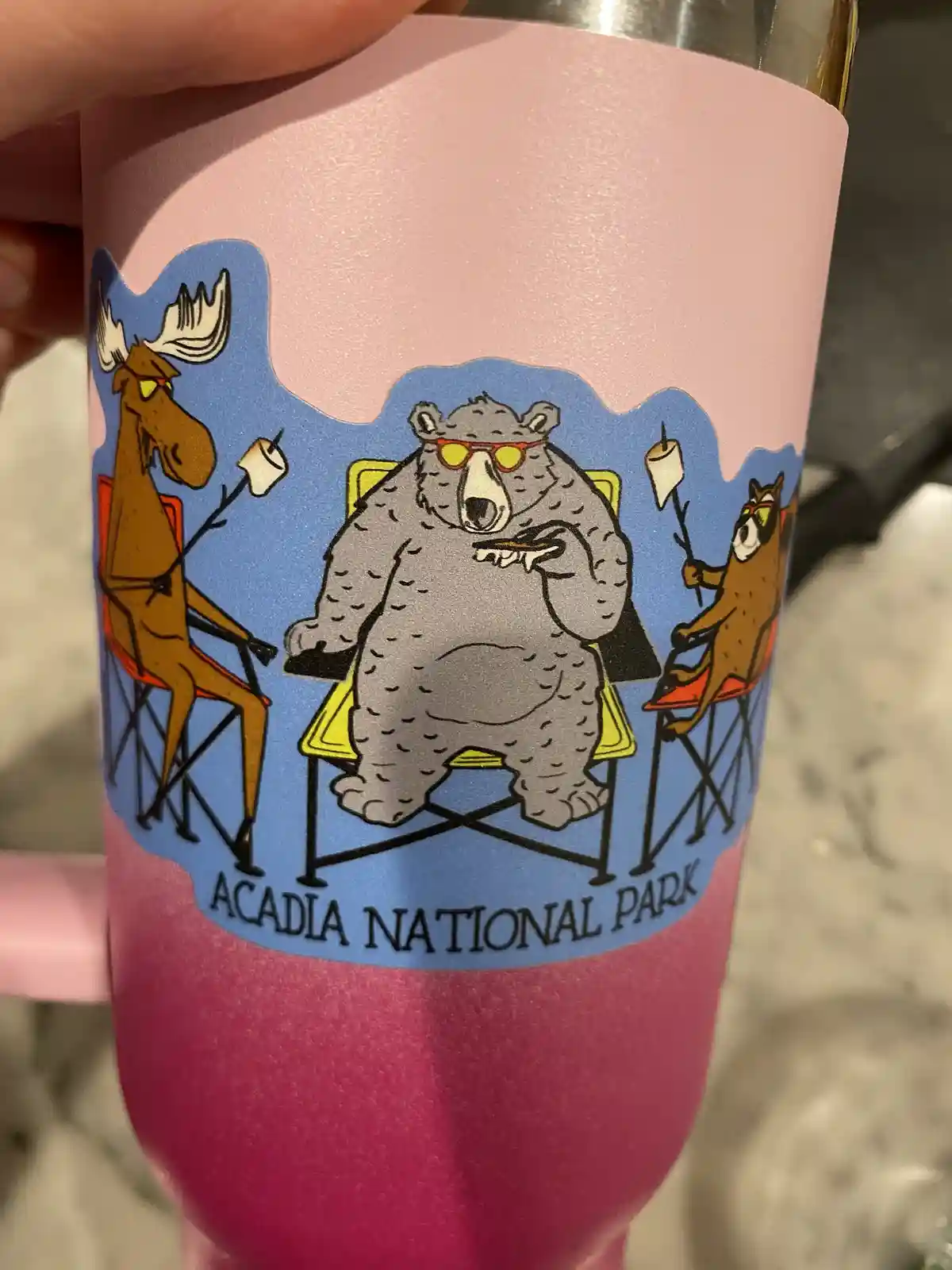 A close-up of a pink insulated tumbler held in a hand, featuring a cartoon sticker of a moose, a bear, and a fox lounging in…