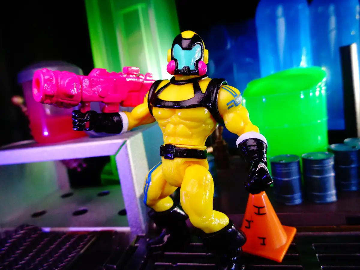 A yellow action figure with a blue face mask and black gloves holds a pink toy gun while standing on a metal grate next to…