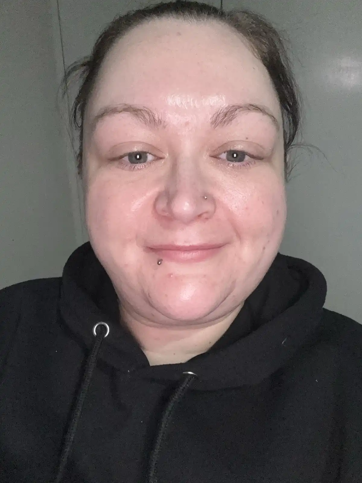 A close-up selfie of a person with dark hair pulled back, wearing a black hoodie, smiling slightly while looking at the…
