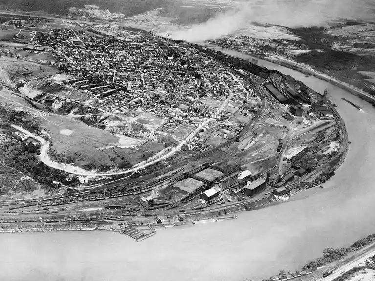 A black-and-white aerial photograph shows a sprawling industrial town nestled along a wide river bend, with numerous train…