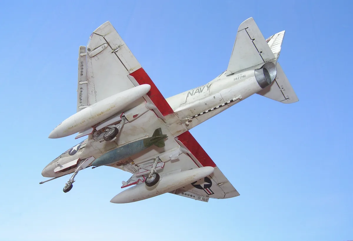 A white and red military jet, identified as a Grumman A-6 Intruder with 'NAVY' and '147741' markings, is shown in flight…