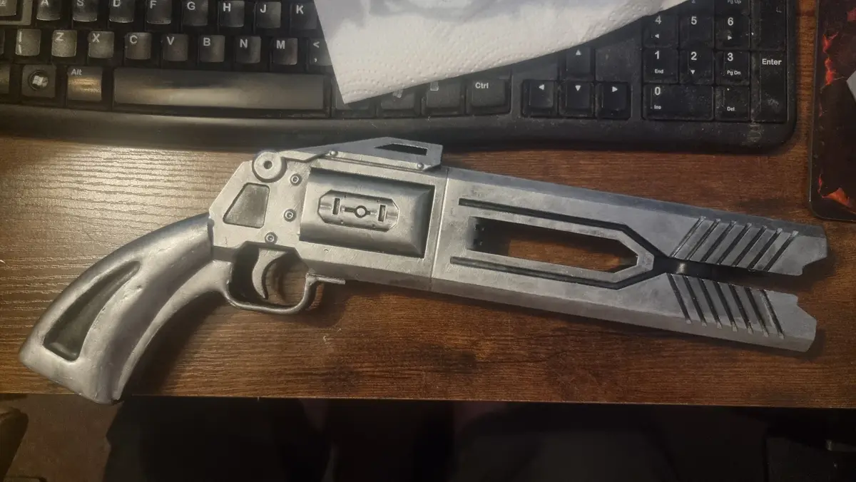 A silver, 3d-printed replica of a futuristic revolver lies on a wooden desk, positioned in front of a black computer…