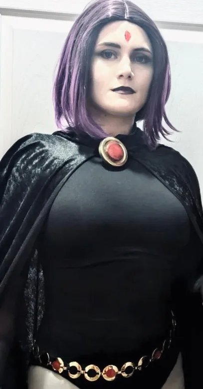 A person in a cosplay costume resembling the character Raven from Teen Titans, wearing a black outfit with a cape, a red gem…