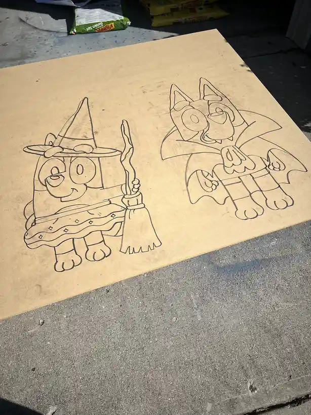 A large piece of cardboard on a concrete surface has two outlined drawings of the cartoon characters Bluey and Bingo dressed…