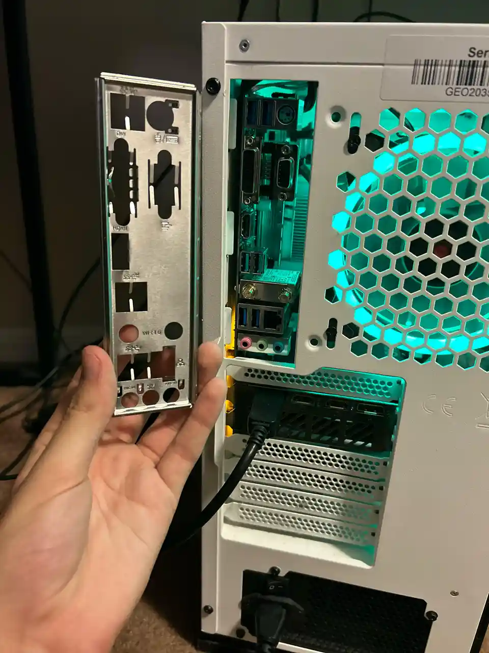 A hand holds up the back panel of a computer's I/O shield, which has cutouts for various ports like HDMI, USB, and Ethernet…