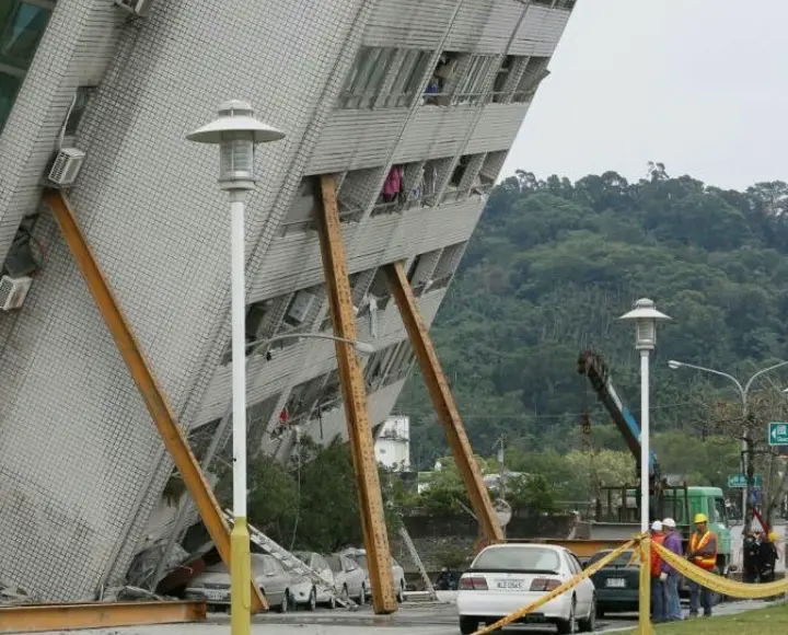 a photograph shows a multi-story building severely tilted sideways, propped up by large wooden beams for structural support…