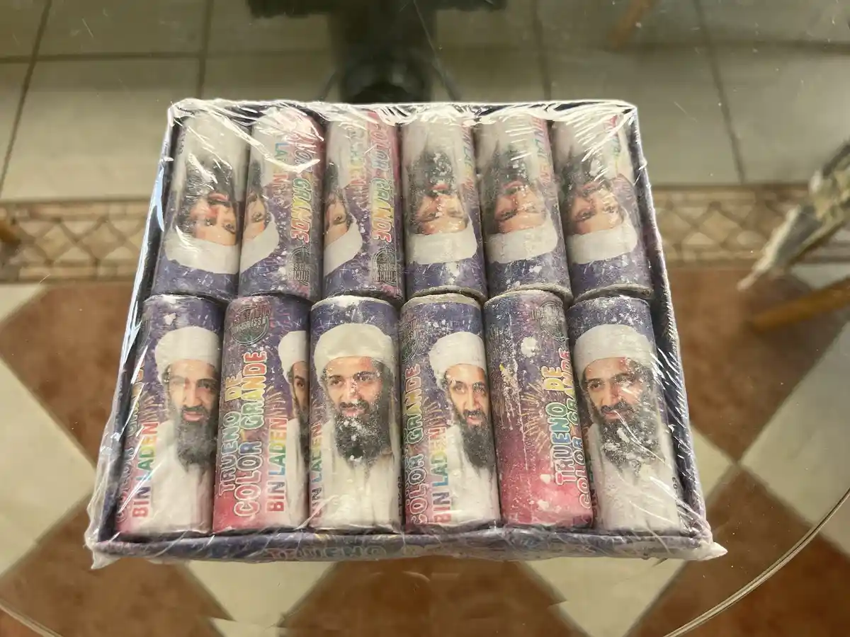 A box of colorful firecrackers, each labeled 'TRUENO DE COLOR GRANDE' and featuring a printed image of Osama bin Laden, is…
