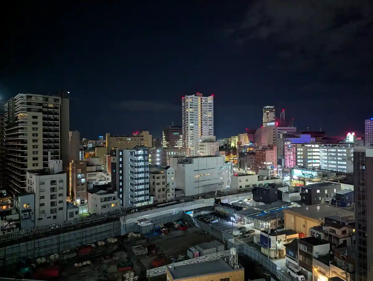 A nighttime cityscape photo of a dense urban area with illuminated high-rise buildings, streets, and a construction site in…