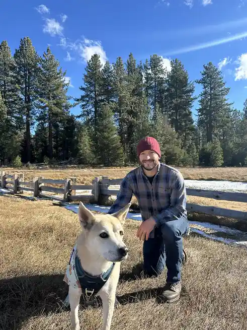 A man in a red beanie and plaid shirt kneels in a grassy field beside a light-colored dog wearing a patterned vest. Behind…