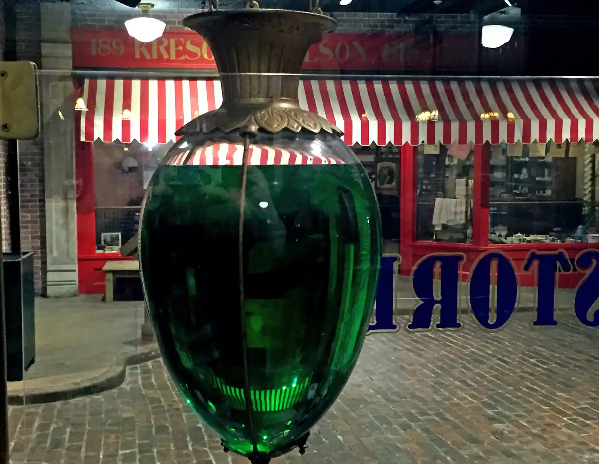 A large, ornate green glass lantern with a brass top hangs in the foreground, partially obscuring a view of a red storefront…