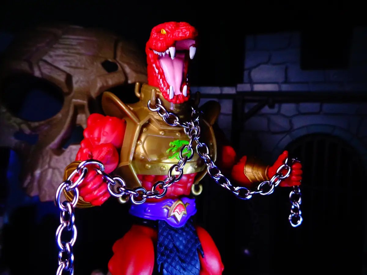 A red, reptilian action figure with a gold chest plate and blue skirt is posed with a large silver chain wrapped around its…