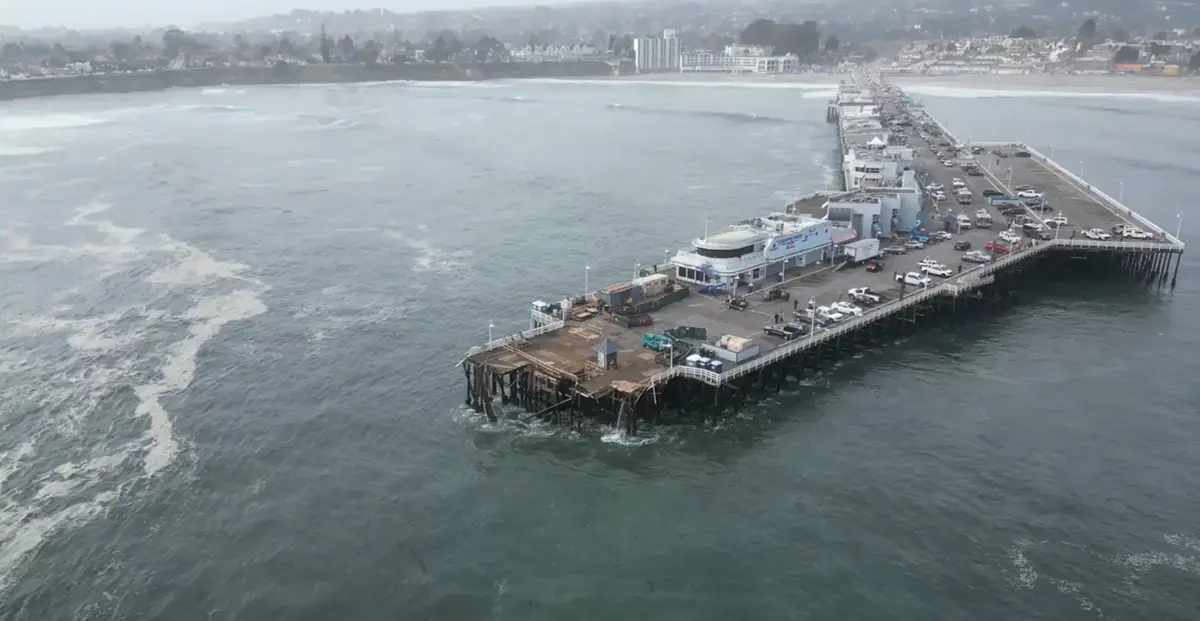 An aerial view shows a long pier extending into the ocean, with visible structural damage and water rushing around its…