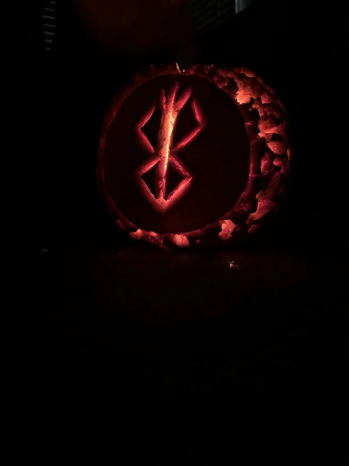 A carved pumpkin illuminated from within in a dark room, featuring a glowing red symbol resembling the Bluetooth logo. The…