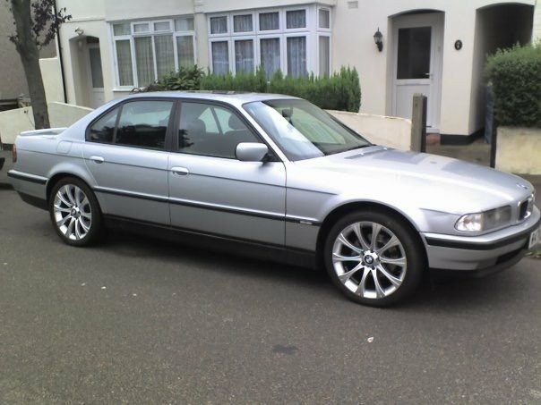 a silver bmw 7 series sedan is parked on a residential street in front of a white house with bay windows and green bushes…