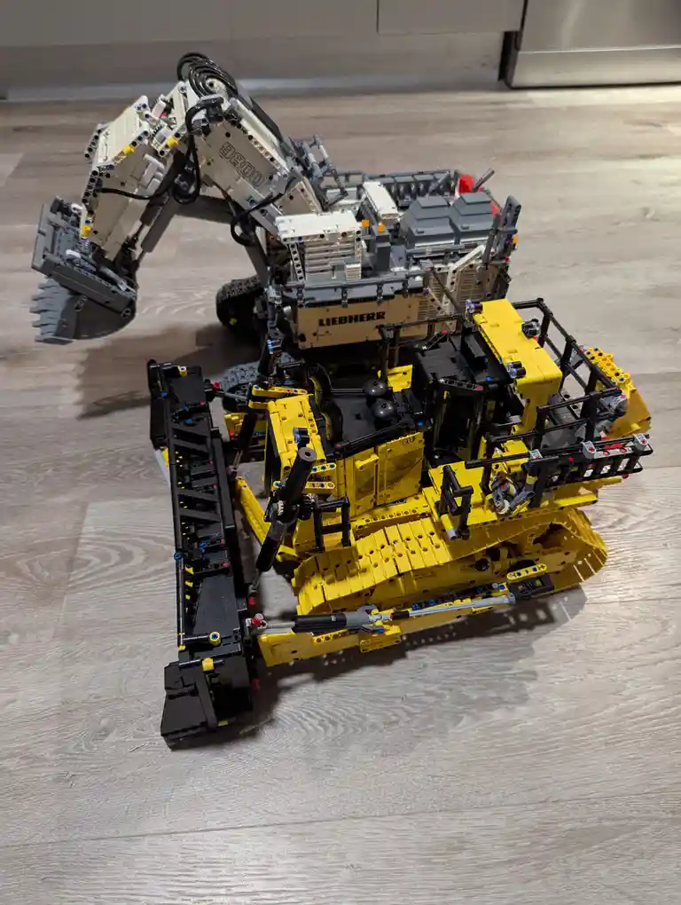 A detailed Lego Technic model of a Liebherr 9800 excavator is shown on a light wood floor, with its articulated arm raised…