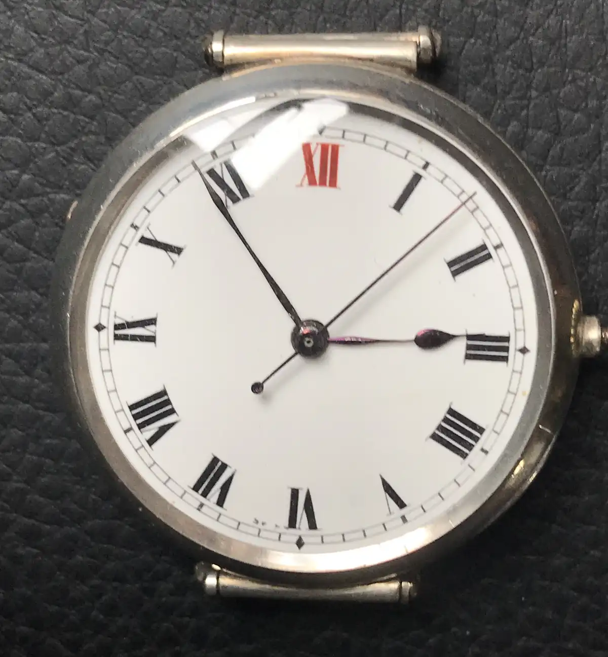A close-up of an antique silver pocket watch with a white face and black Roman numerals, resting on dark textured leather…