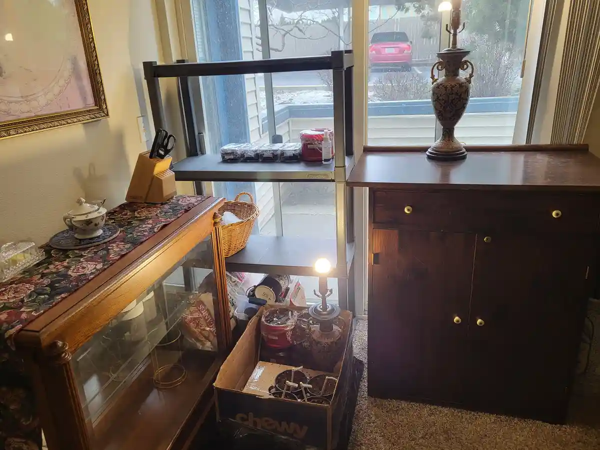 A room corner shows a wooden cabinet with a decorative lamp on top, next to a glass display case with a teapot and a knife…