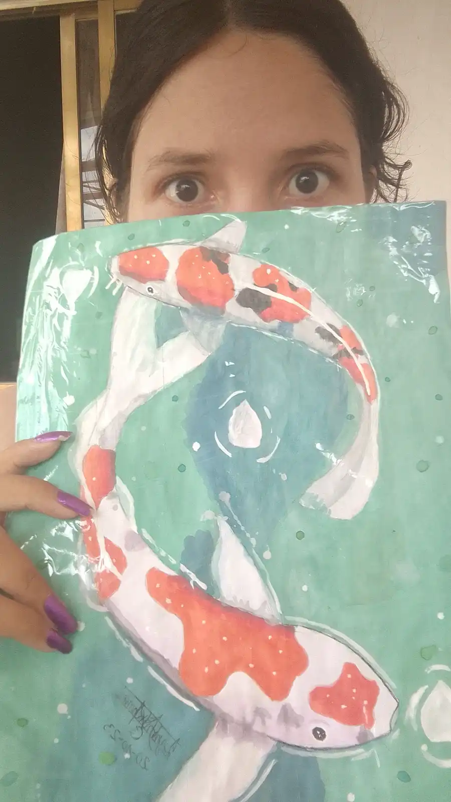 A person with dark hair and wide eyes holds up a painting of two koi fish, covering their face except for their eyes. The…