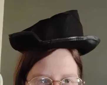 A person with glasses and brown hair wears a black, oddly shaped hat that looks like a distorted, flattened fedora. The hat…