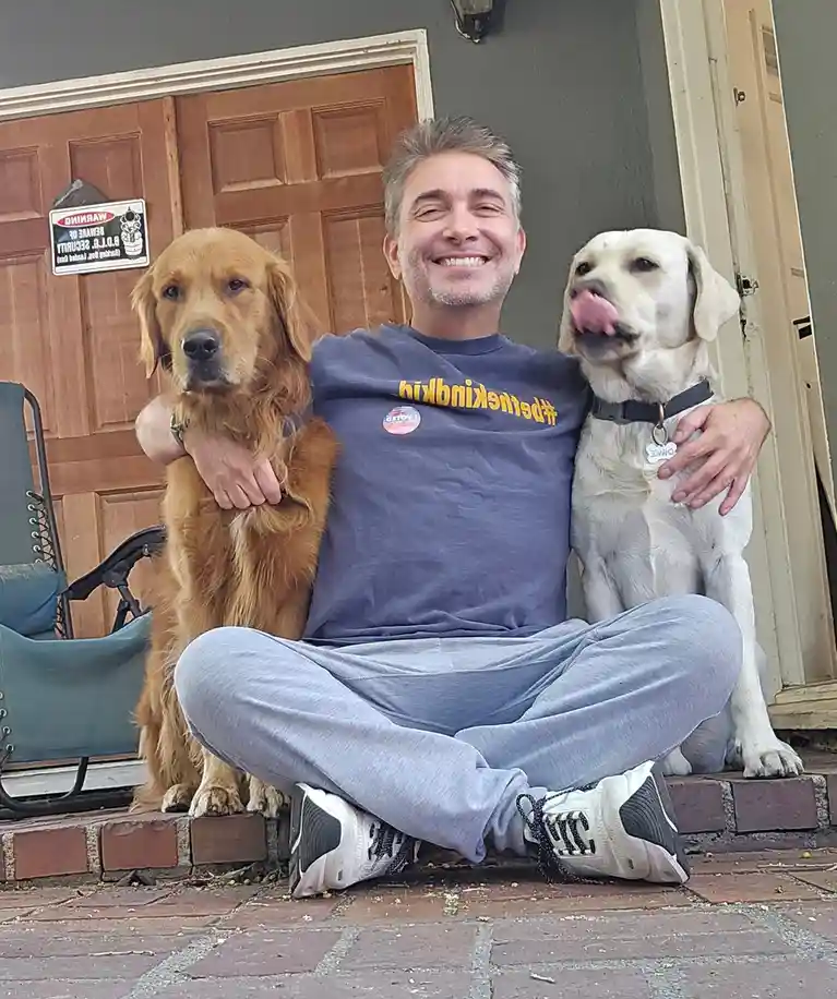 A man sits cross-legged on a brick porch with two dogs, one golden retriever and one yellow lab, both being hugged. He wears…