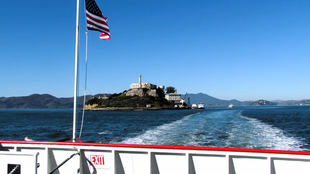 A photo taken from a boat looking toward Alcatraz Island, with an American flag flying on the left and an 'EXIT' sign…