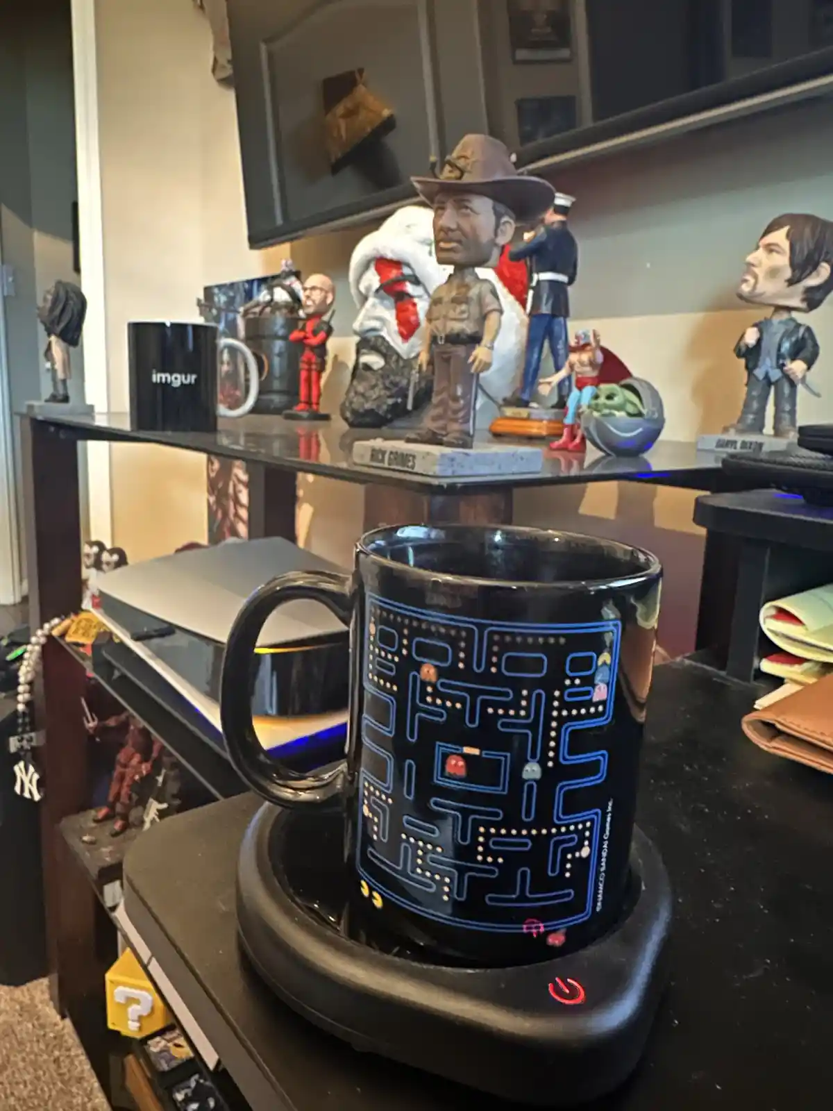 A black mug with a Pac-Man maze design sits on a heated coaster, surrounded by pop culture figurines including Rick Grimes…