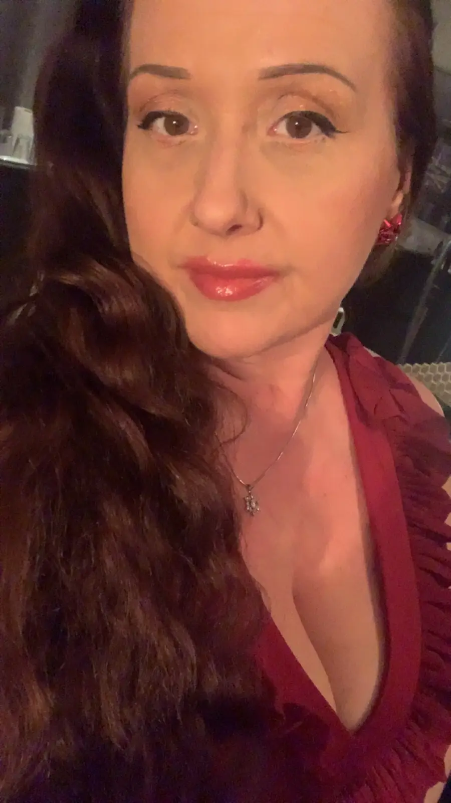 A close-up selfie of a woman with long dark wavy hair, wearing a red ruffled top, red earrings, and a necklace, looking…