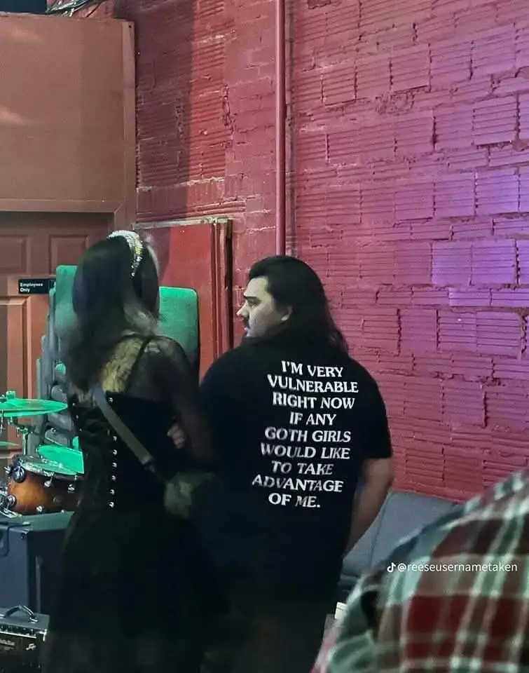 A man with long dark hair, seen from behind, wears a black t-shirt with white text that reads 'I'M VERY VULNERABLE RIGHT NOW…