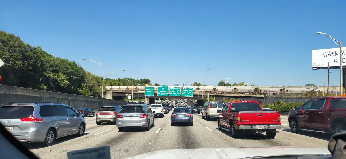 A photo taken from inside a car shows heavy traffic on a multi-lane highway under clear blue skies, with green highway signs…